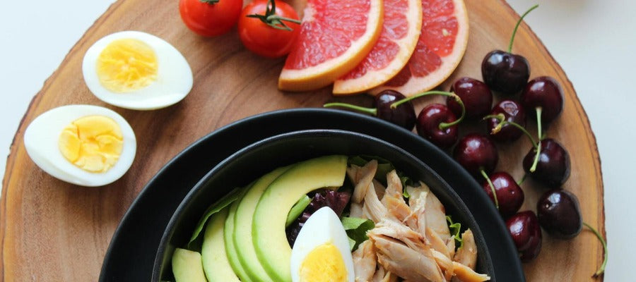 round wooden platter with sliced egg, tomatoes, grapefruit, sour cherries and bowl with avocado and other healthy food