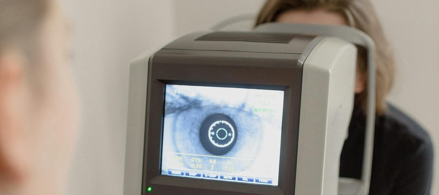 ojo visto en el monitor de un dispositivo de examen ocular con una mujer sentada detrás de él en el fondo y la cara borrosa del oftalmólogo en primer plano