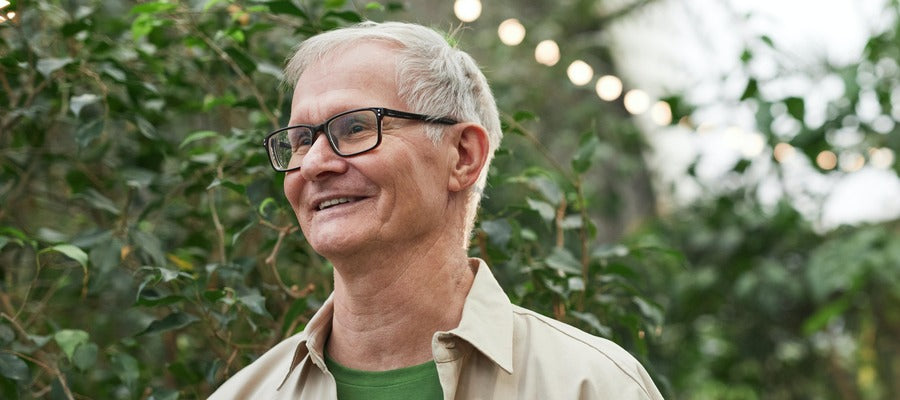 man with white hair and black glasses looking away and smiling against leafy green background