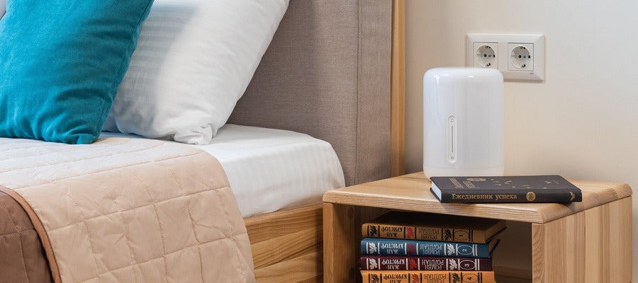 white humidifier lying on nightstand next to made bed under shelf with books and with wall sockets behind it
