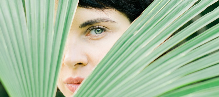 woman with bright green eye and dark hair hiding one eye and half of her face before green fan-like plant