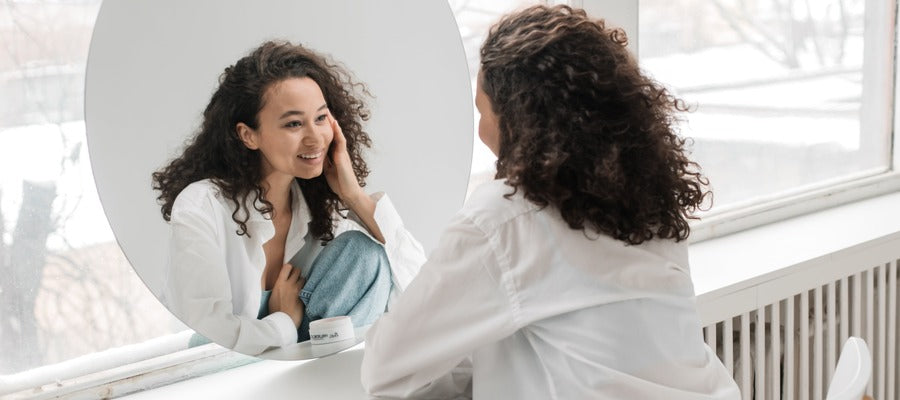 calm and smiling woman looking at herself in a round mirror resting on the windowsill