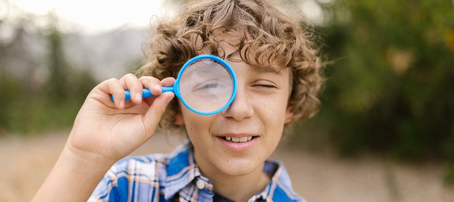 young boy in shirt holding magnifying glass over one eye outdoors with blurry field and trees in the background