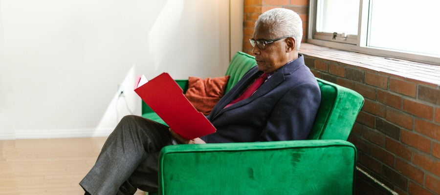 man with glasses reading a red folder in a green couch against a brick background