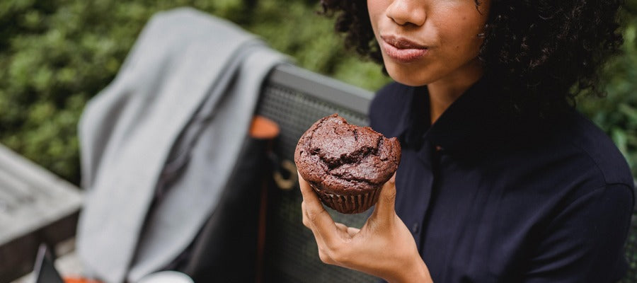 Mujer con peinado afro llevándose un pastelito de chocolate a la boca mientras está sentada en un banco afuera