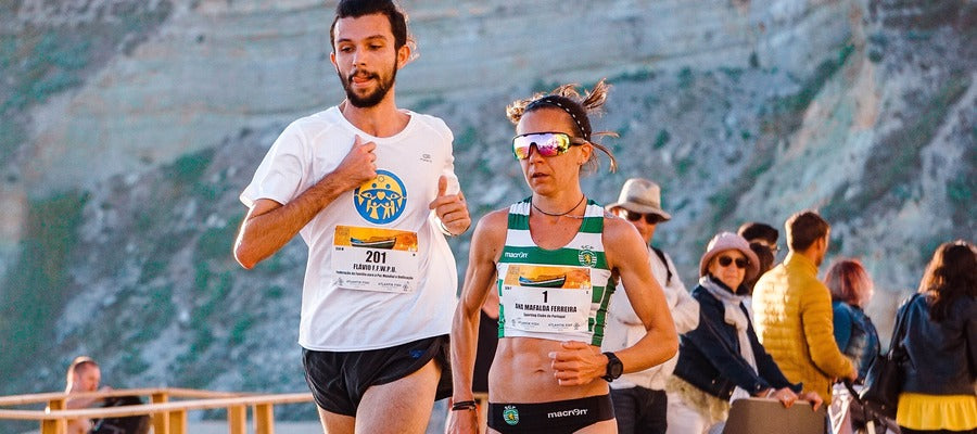 female runner with sunglasses outdoors during race with male runner in white shirt without sunglasses and people watching them in the background