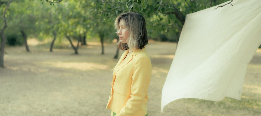 woman in yellow shirt standing with eyes closed outside on windy day with windblown sheet before her on a line and green trees in the background