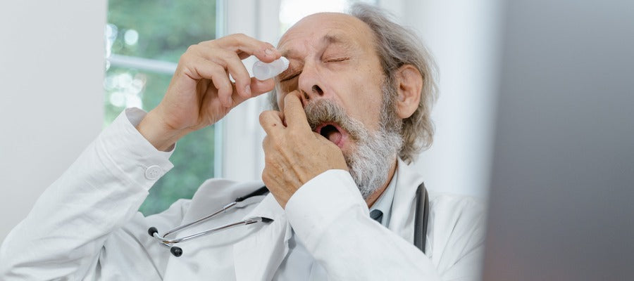 old doctor with gray hair and beard applying eye drops in one eye while sitting at his desk with green window in the background