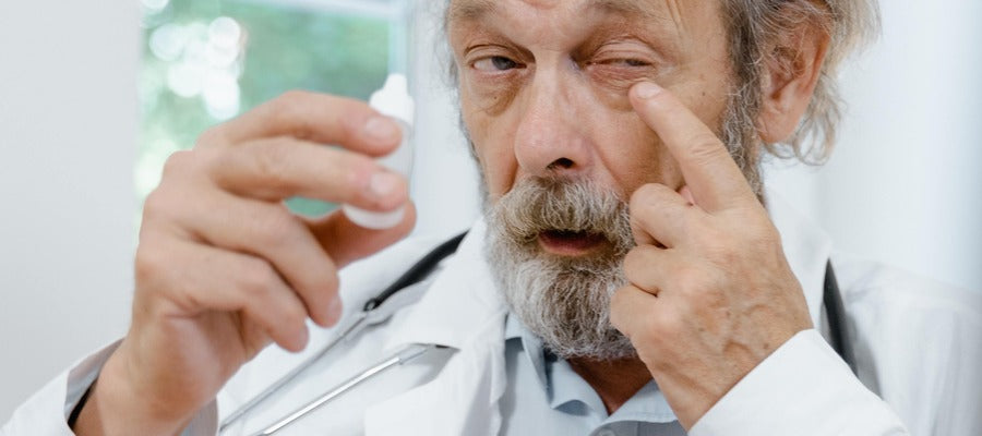 old doctor in a white coat holding eye drops bottle in one hand while touching eyelid with one finger
