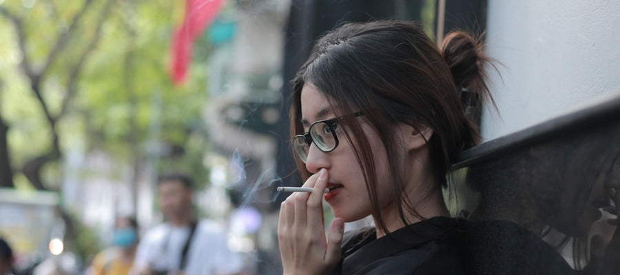 Mujer con cabello oscuro y gafas fumando un cigarrillo afuera vista de perfil
