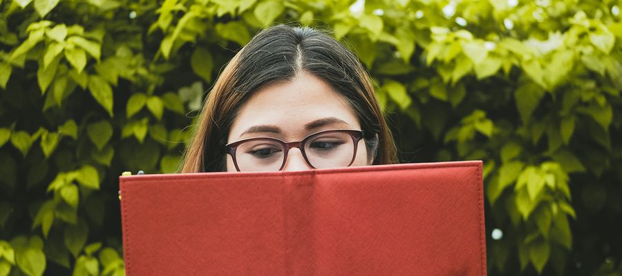 woman with eyeglasses reading from a red book with greenery in the background