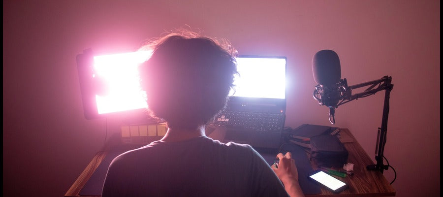 man seen from behind in a darkened room with glowing computer screens and microphone before him