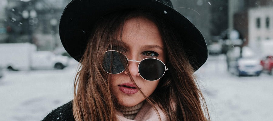 Mujer con sombrero y gafas de sol redondas contra el fondo urbano cubierto de nieve