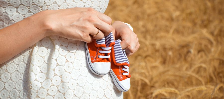 Mujer embarazada con vestido blanco sosteniendo zapatos de bebé rojos