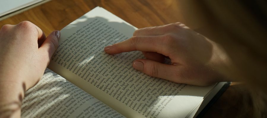 Persona leyendo un libro bajo una luz tenue con el dedo en la página derecha visto por encima del hombro
