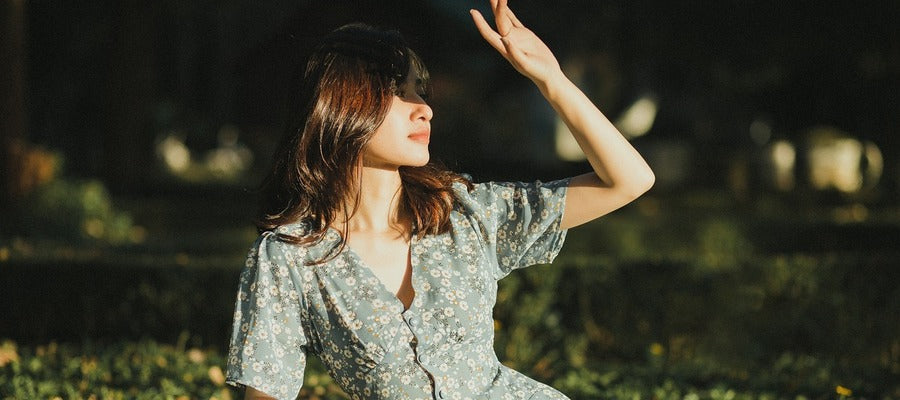 woman in a light summer dress sitting on grass and shielding her eyes from the bright sun