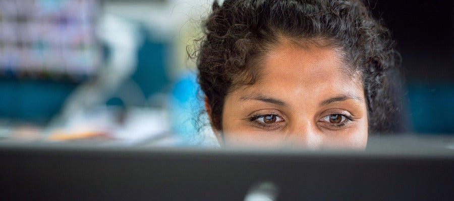 woman behind computer screen looking down at it with tired eyes against blurry background