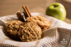 Fresh baked, crumble covered apple cinnamon muffins made with cinnamon and clove essential oils in a basket lined with a towel and an apple in the background.