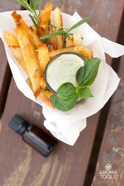 A basket of rosemary baked potato fries seasoned with rosemary essential oil, a cup of creamy basil dip, and sprigs of rosemary and basil.