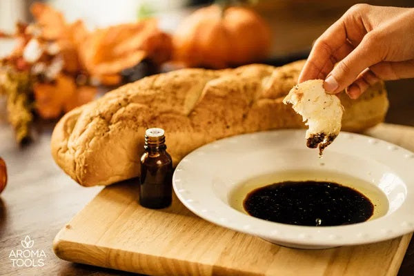 A hand dipping a slice of French bread into a bowl of essential oil-infused Italian dipping oil sitting on a wooden cutting board with a loaf of French bread.
