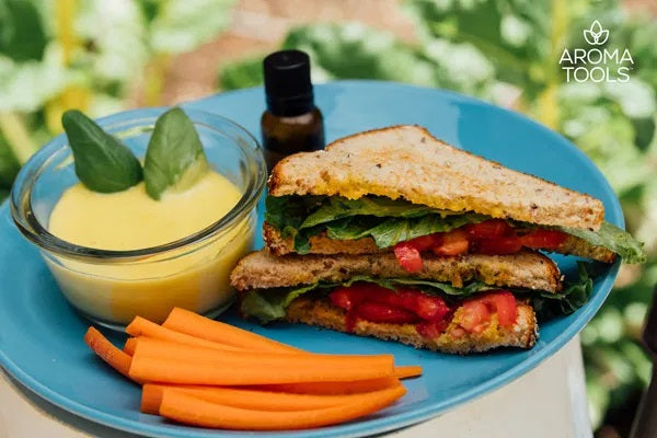 A veggie sandwich on wheat bread with carrot sticks and a glass bowl with a generous serving of our homemade mayonnaise flavored with essential oils sitting on a plate outside.