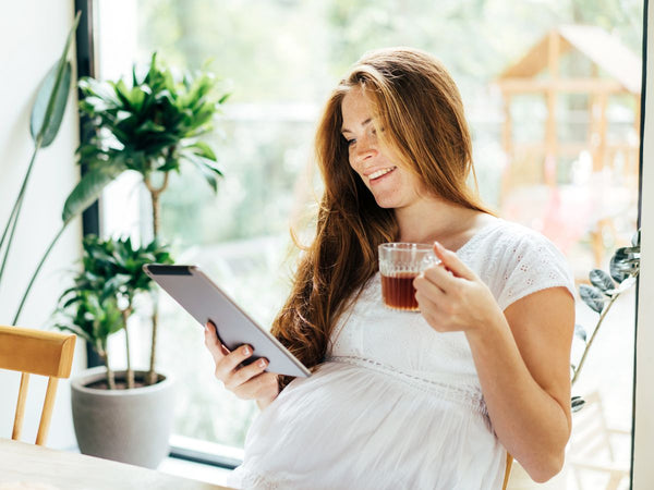 pregnant woman sitting at a table drinking coffee and looking at a tablet device