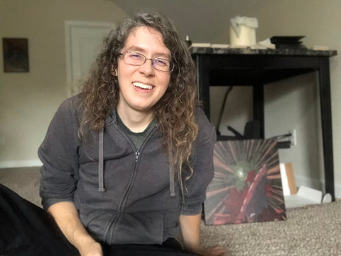 A person with long curly brown hair and glasses, in a gray zip up hoodie and black jeans is sitting on the floor smiling.  A table is behind her, and a canvas print is on the floor.