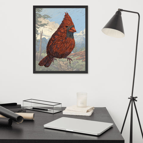 A framed art print of a hand drawn cardinal in front of a faded background.  The print is hanging on a white wall over a cluttered desk with a lamp to the right.