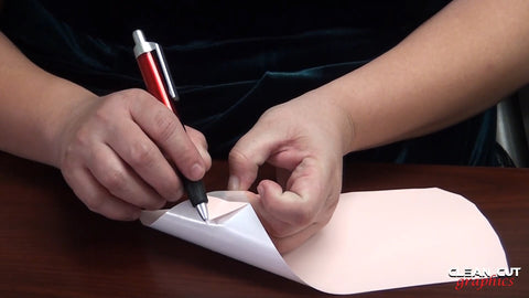 A close-up showing two hands, one is holding the carrier sheet in place while the other is peeling the vinyl with a weeding tool from the corner of the design. 