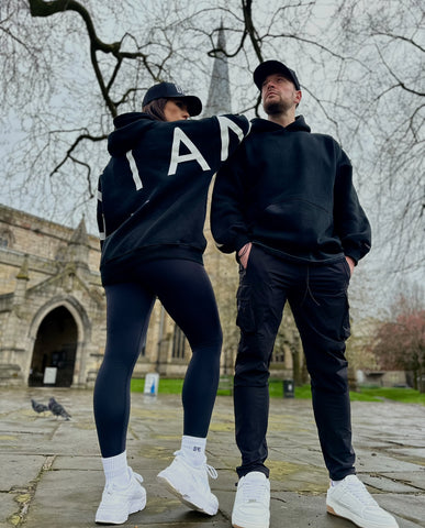 A guy and a girl stood posing in front of a church on a cloudy day, both dressed in the amazing Oversized DFNT. Eternity Hoodies.  The Hoodies have a plain front with a big DEFIANT lettering across the arms and back, a subtle DFNT. tag on the left side and then embroidery stating Proudly Refusing To Obey Authority under the large lettering as well as The Defiant Co embroidered to the left sleeve cuff.  The hoodies are black with white logos.