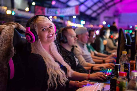 Ein blondes Mädchen mit einem roten Headset sitzt vor einem Computer und lächelt in die Kamera. Dahinter sitzen weitere Computerspieler in einer Reihe