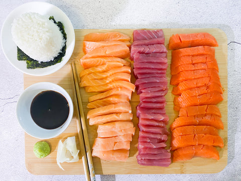 Bessie Box sashimi-grade Atlantic salmon, ahi tuna, and sockeye salmon sliced and served as sashimi with soy sauce, rice, wasabi and ginger on a bamboo board.