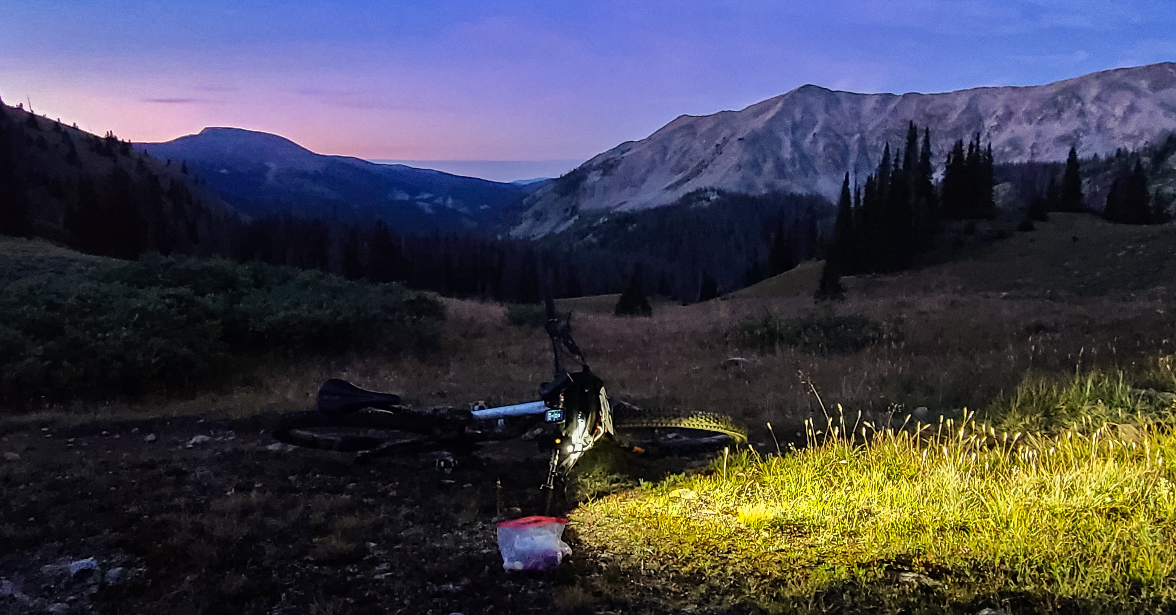 Mountain landscape photo with sun setting in background and bike laying on ground in foreground