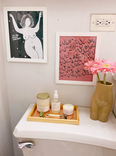 two framed art prints hanging on a wall above a shelf that has a tray holding a candle, room spray, and lotion; adjacent to the tray on the self is a feminine vase with pink flowers