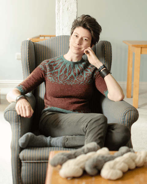 Marina sits in an armchair, chin on hand, wearing a hand-knitted colourwork yoked jumper in teal and brown.