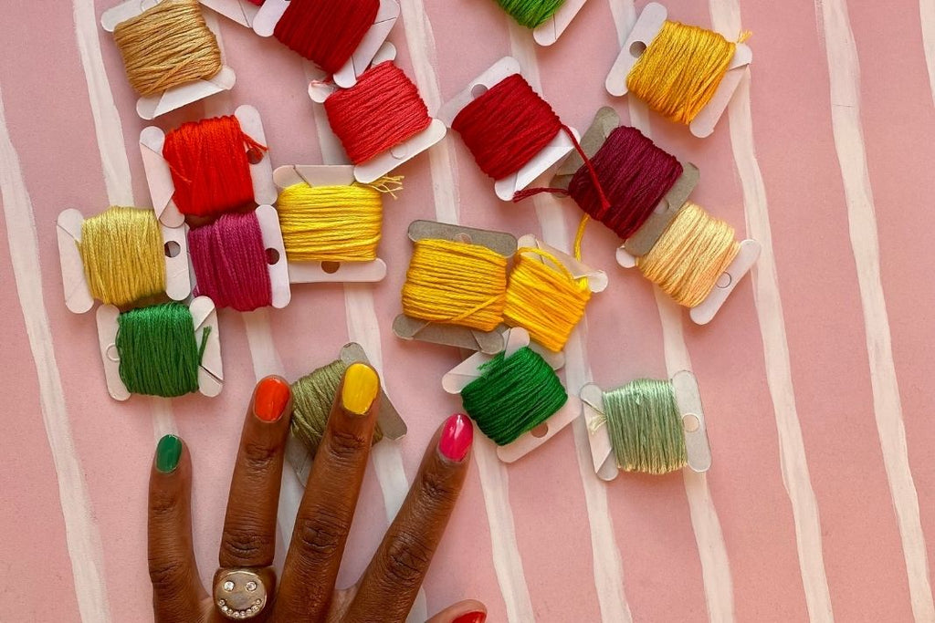 Embroidery threads in red, yellow, maroon and greens, with a hand with matching nail colours beneath them on a pink striped surface.