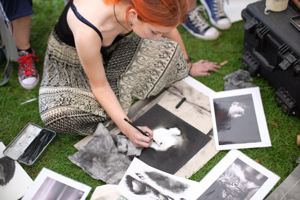 Girl sitting on a grassy floor with charcoal drawings surrounding her as she draws.