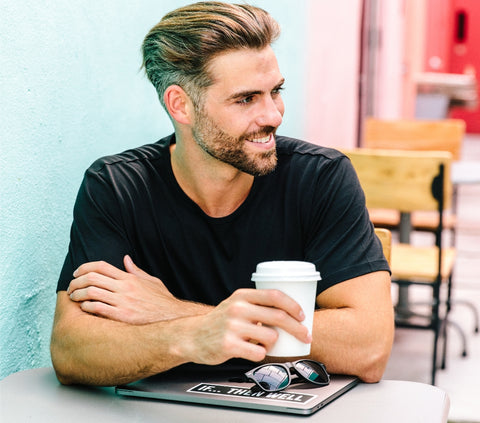 Man in plain black T-shirt sitting and enjoying a coffee with laptop closed in front of him
