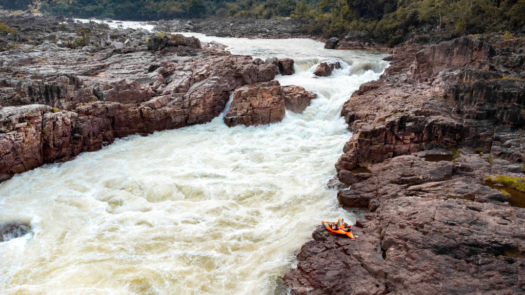 Two kayakers sitting in their boats on the shore while looking upstream at a large rapid on a high volume river.