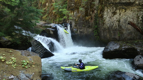 A kayaker sitting in a pool in the river watching another kayaker paddle the waterfall.