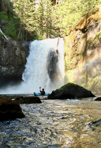 A packrafter paddling across the pool of a river below the waterfall.