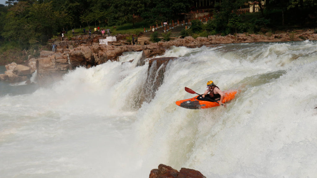 Kayaker paddling off a 15 foot waterfall in front of a small crowd watching in the background.