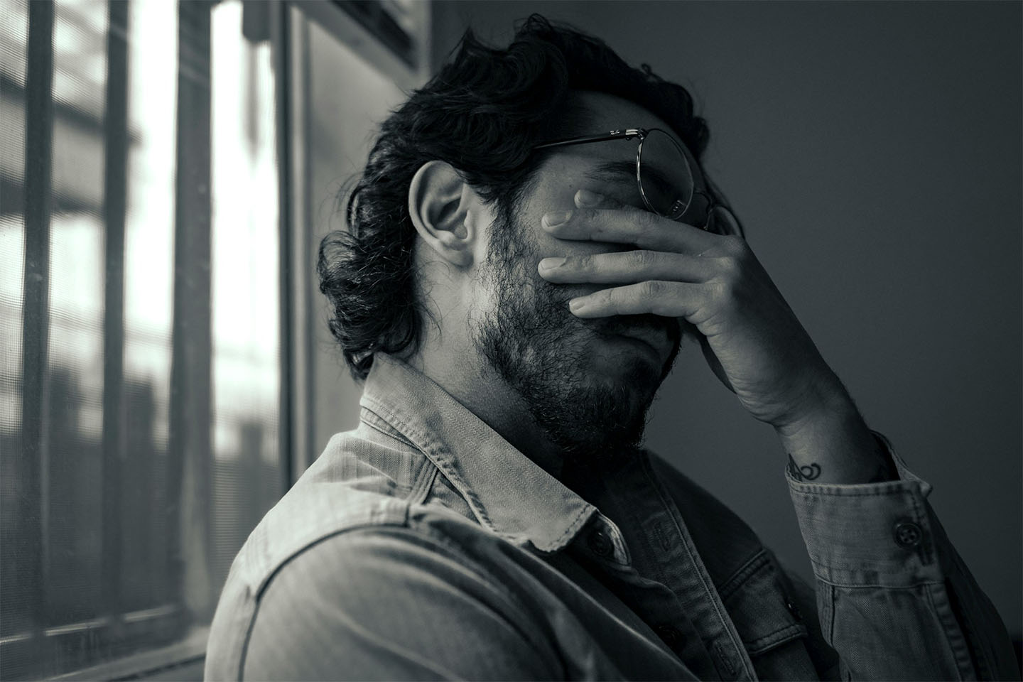 A grayscale photo of a man wearing glasses with his left hand covering his face.