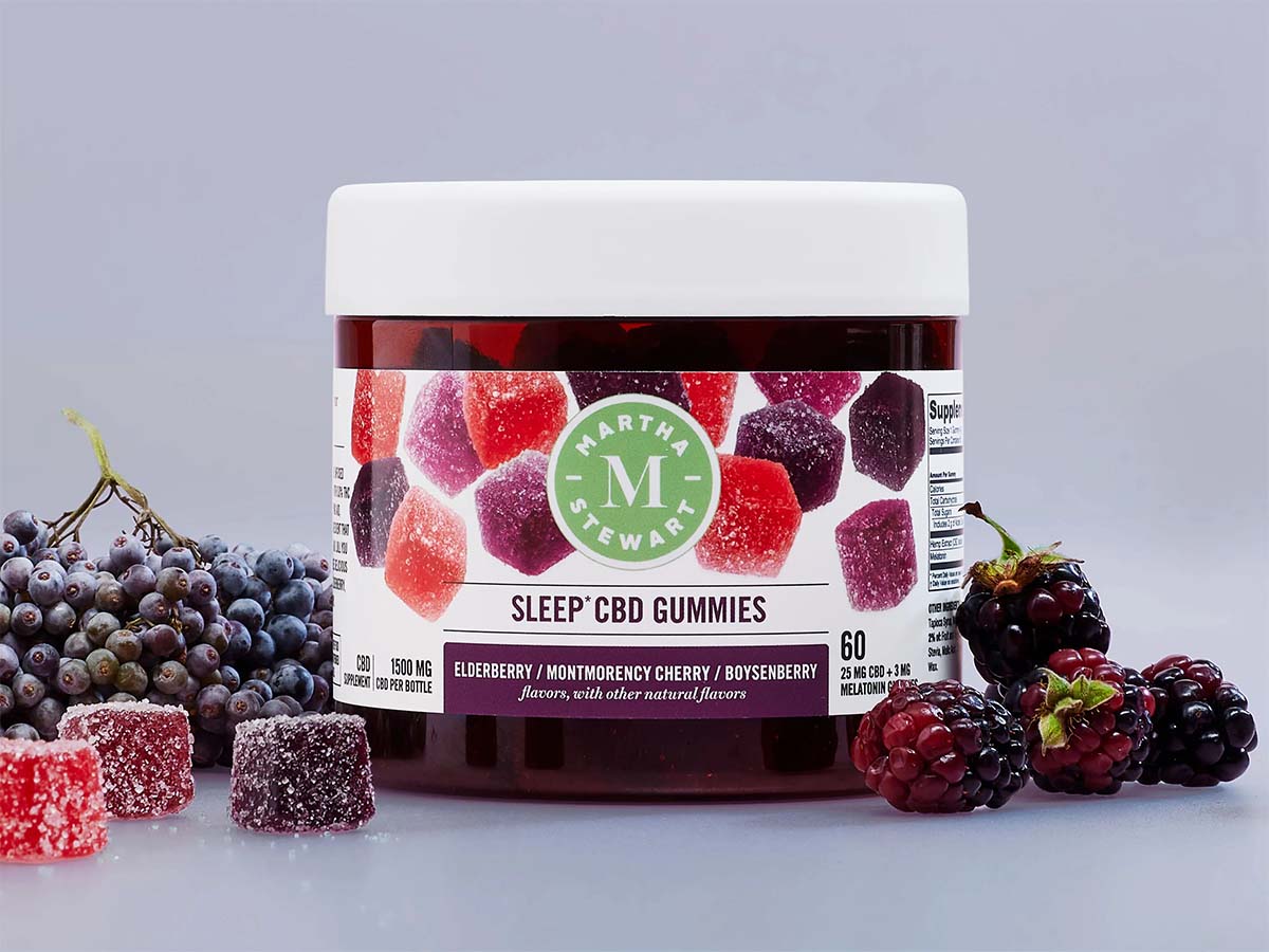 Multicolored gummies and berries around a Martha Stewart Sleep CBD Gummies jar with a white, red, and purple label