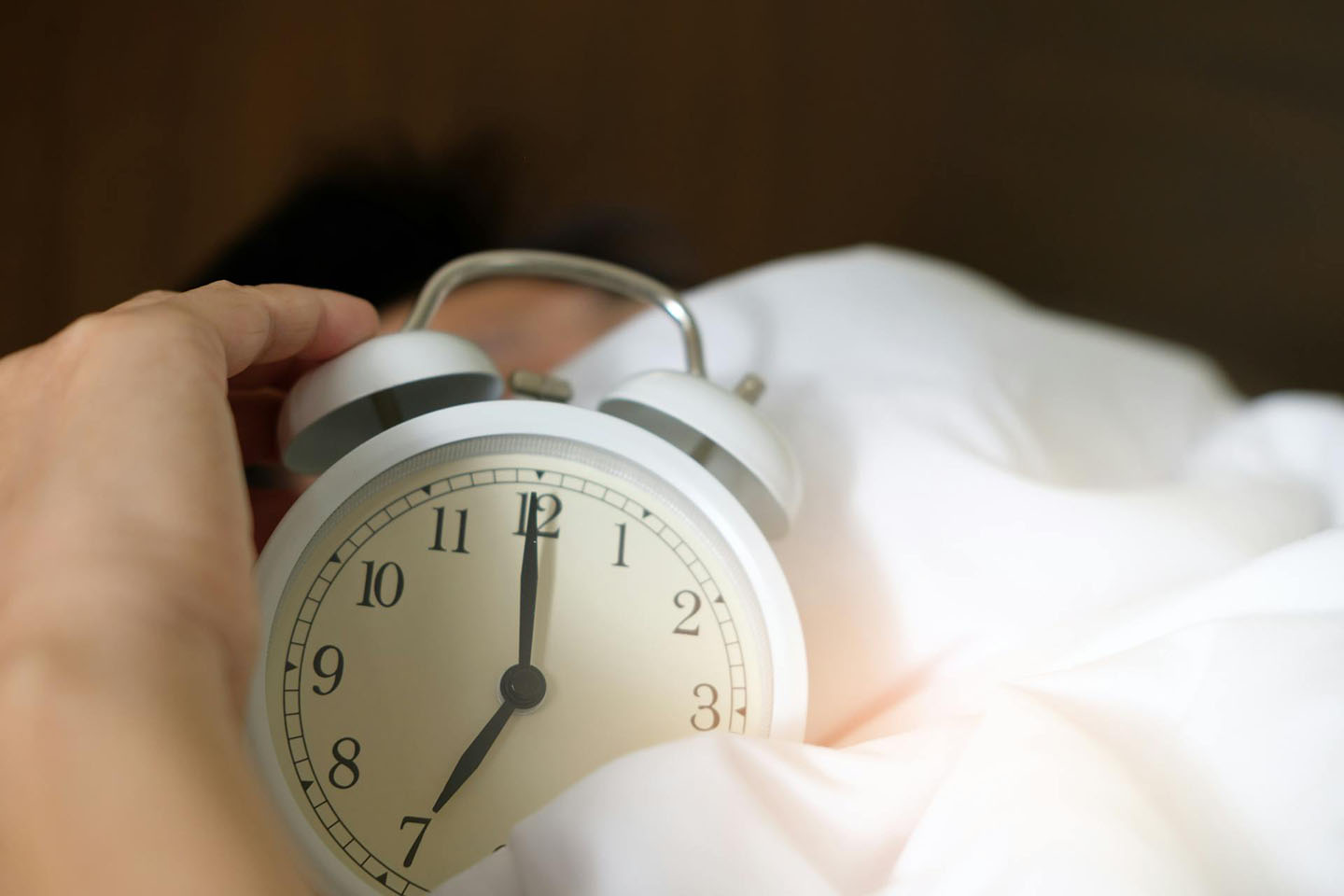 Close-up of person holding a white analog alarm clock in bed