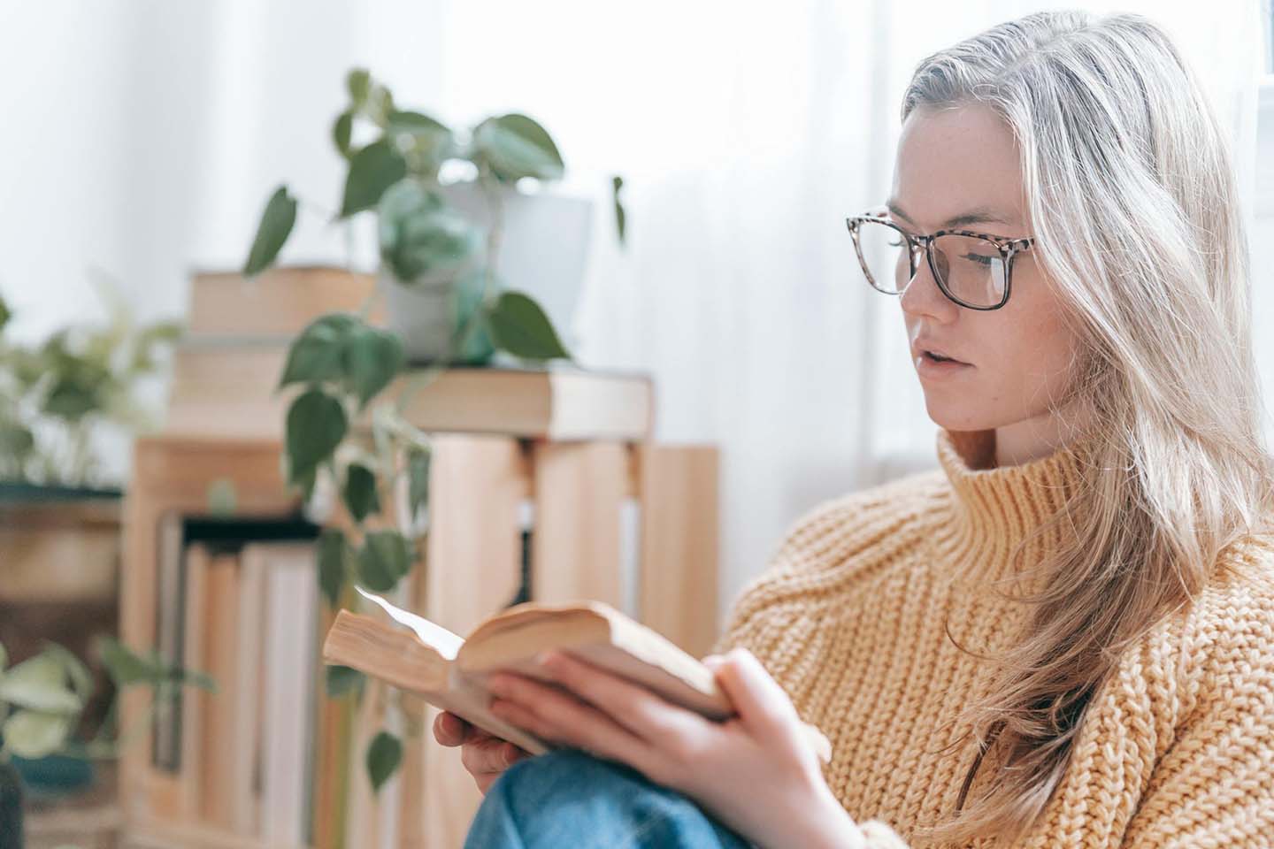 A young woman sitting on the floor while reading a book with a plant hanging in the background