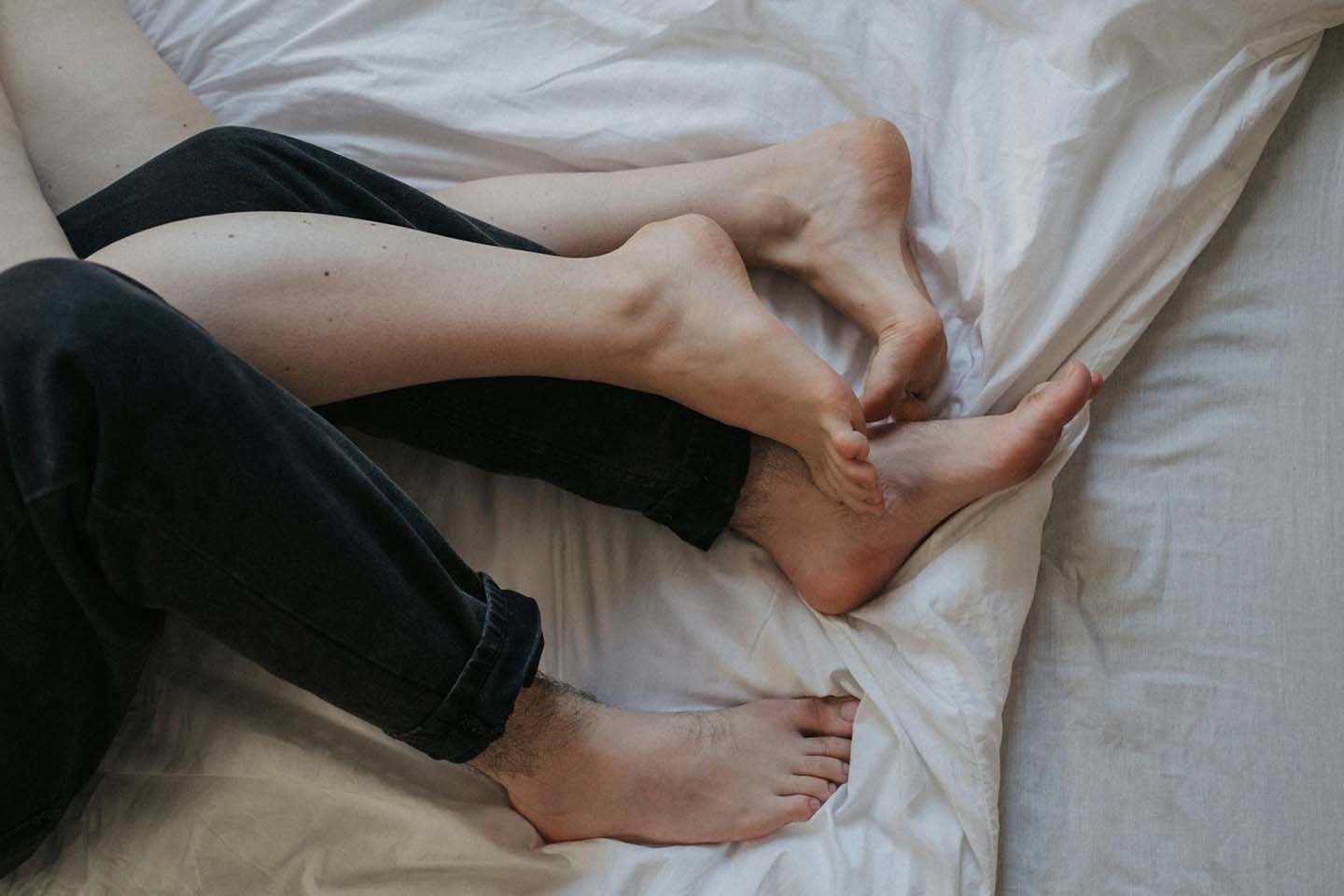 A couple's legs intertwined on a bed coming from the left side of the image. One of them is wearing pants but no socks, the other's legs are bare.
