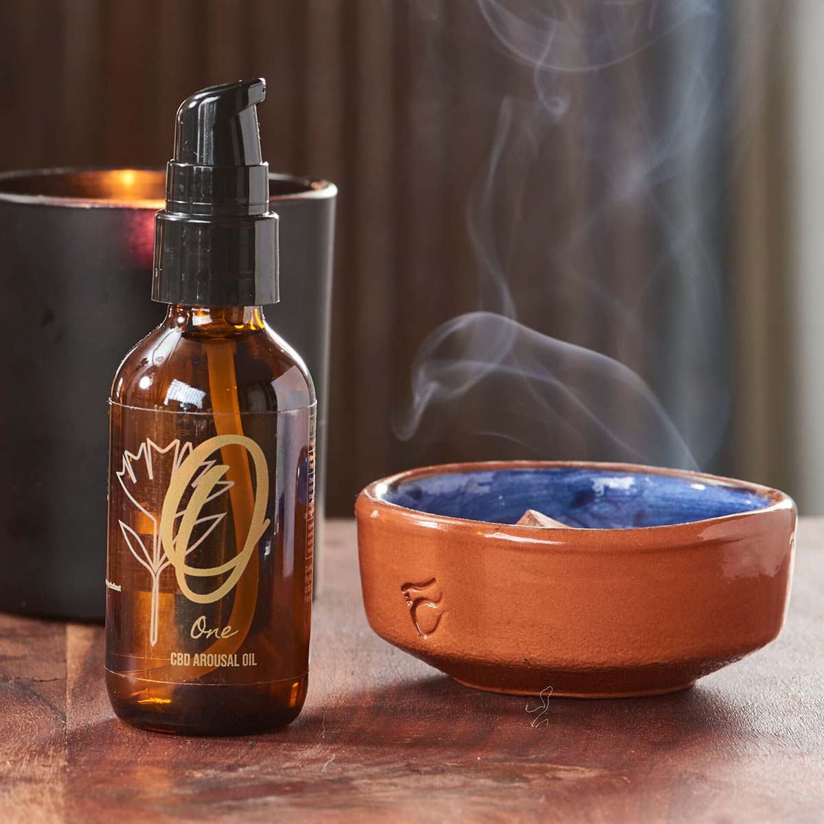 A bottle of Farm to Health's CBD Intimacy oil sits on a wood table in front of a candle and a still-smoking bowl of incense.