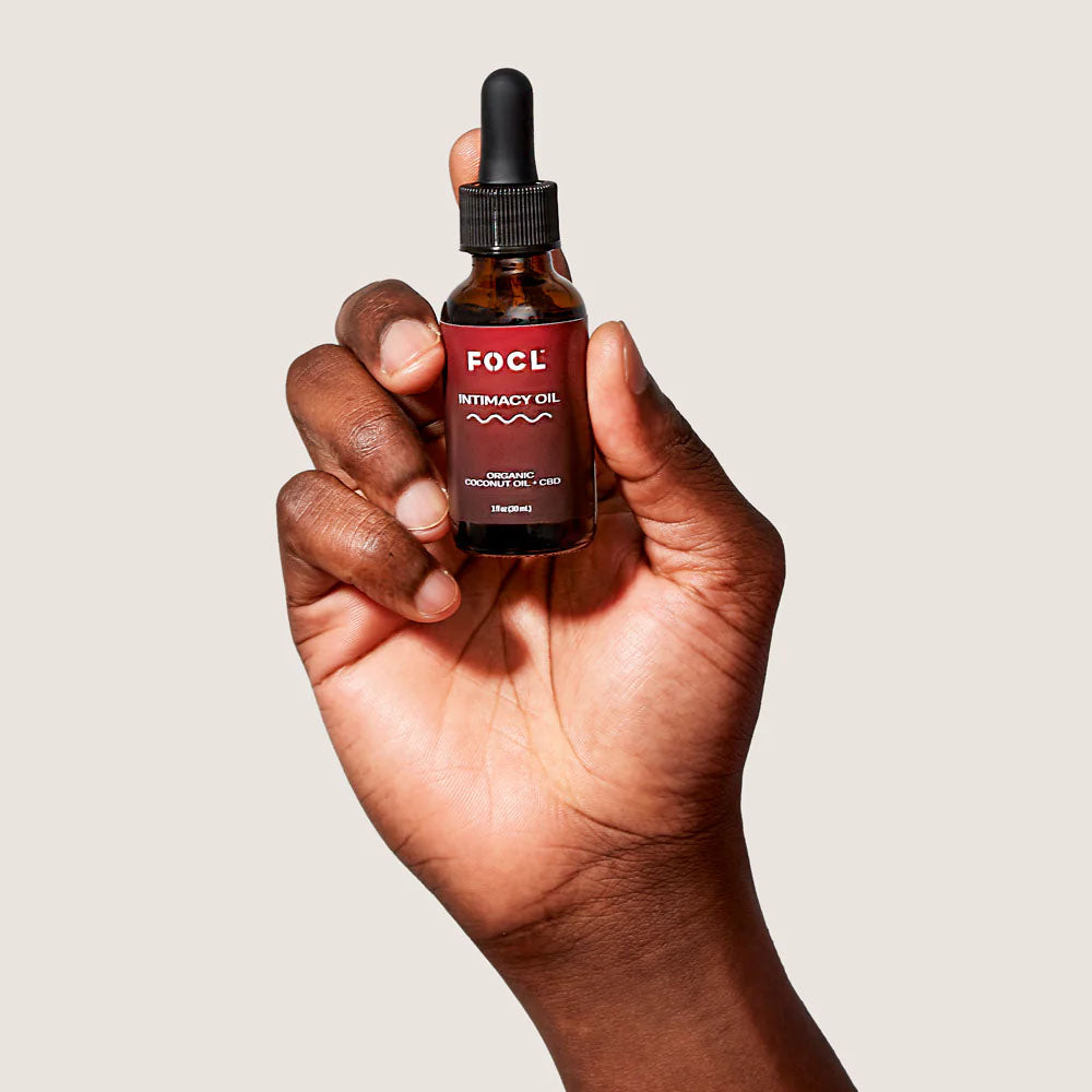 A hand holds a bottle of FOCL Intimacy Oil against a beige background.