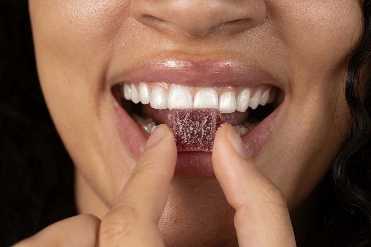 A close up shot as a woman holds a gummy between her index finger and thumb as she bites into it, smiling.